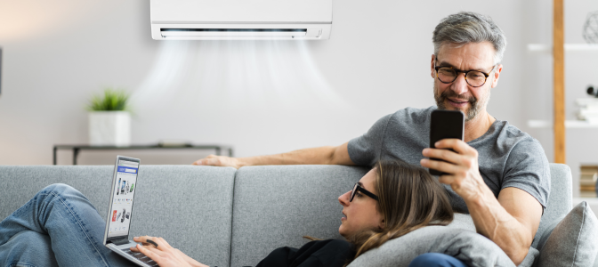 Casal relaxando no sofá com ar-condicionado ligado, aproveitando conforto térmico enquanto usam celular e notebook.