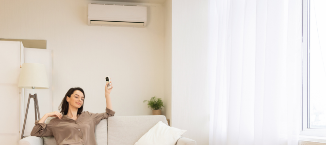 Mulher relaxando em sala climatizada enquanto utiliza controle remoto de ar-condicionado instalado na parede.